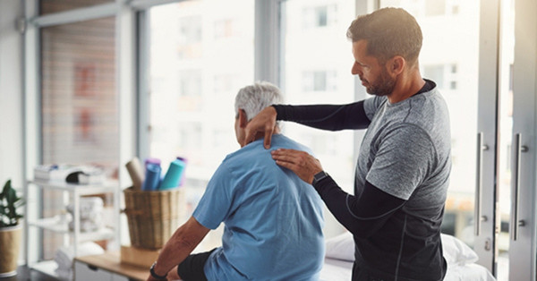 Physiotherapist treating elderly man’s shoulder in bright room.