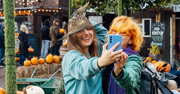 Two friends taking selfie wearing witch hat at pumpkin farm.