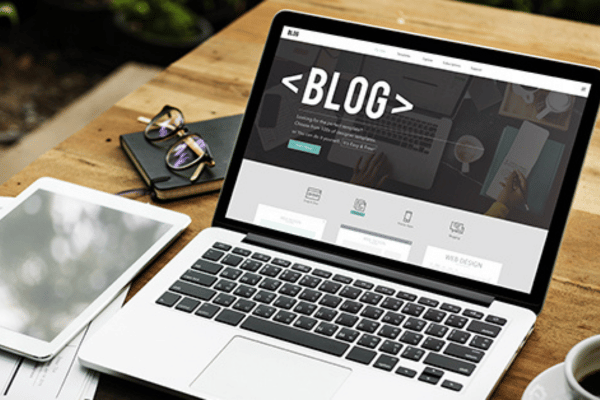 A laptop on a wooden desk displays a blog creation page beside a tablet and papers.