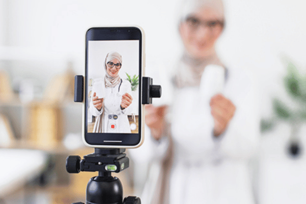 A doctor in a headscarf films a health related video on a smartphone mounted on a tripod.