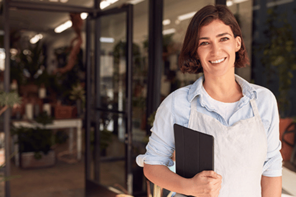 A smiling healthcare practice owner stands at her clinic entrance holding a digital tablet.